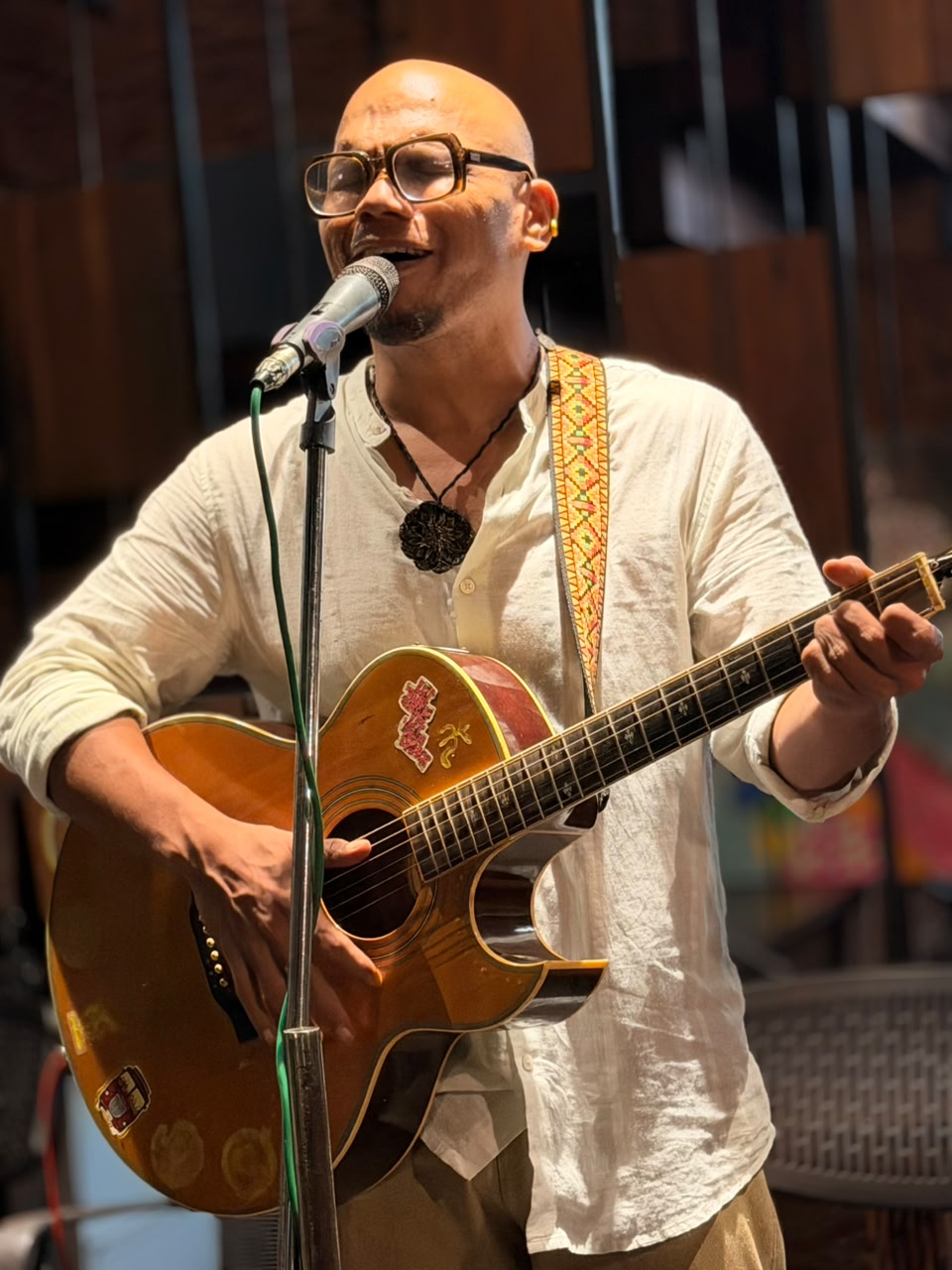 Saji Oorali singing into a microphone with an acoustic guitar, in daylight.