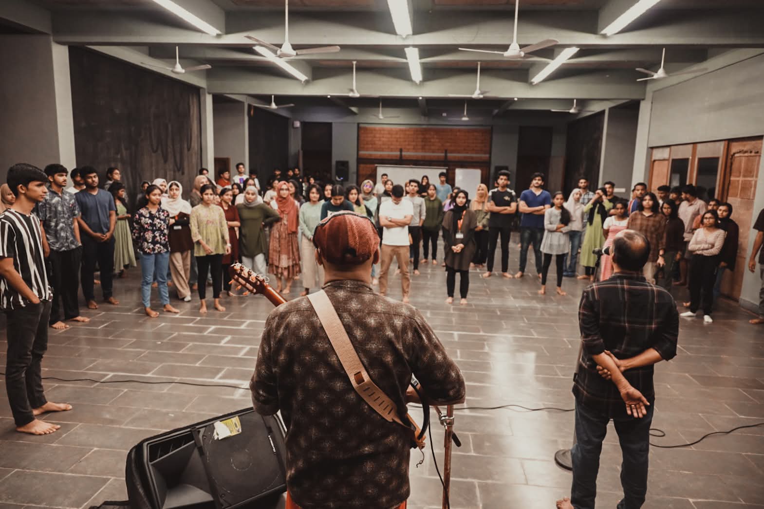 A large group of students stands in a wide circle in a college hall, barefoot, facing a facilitator with a guitar at the centre.