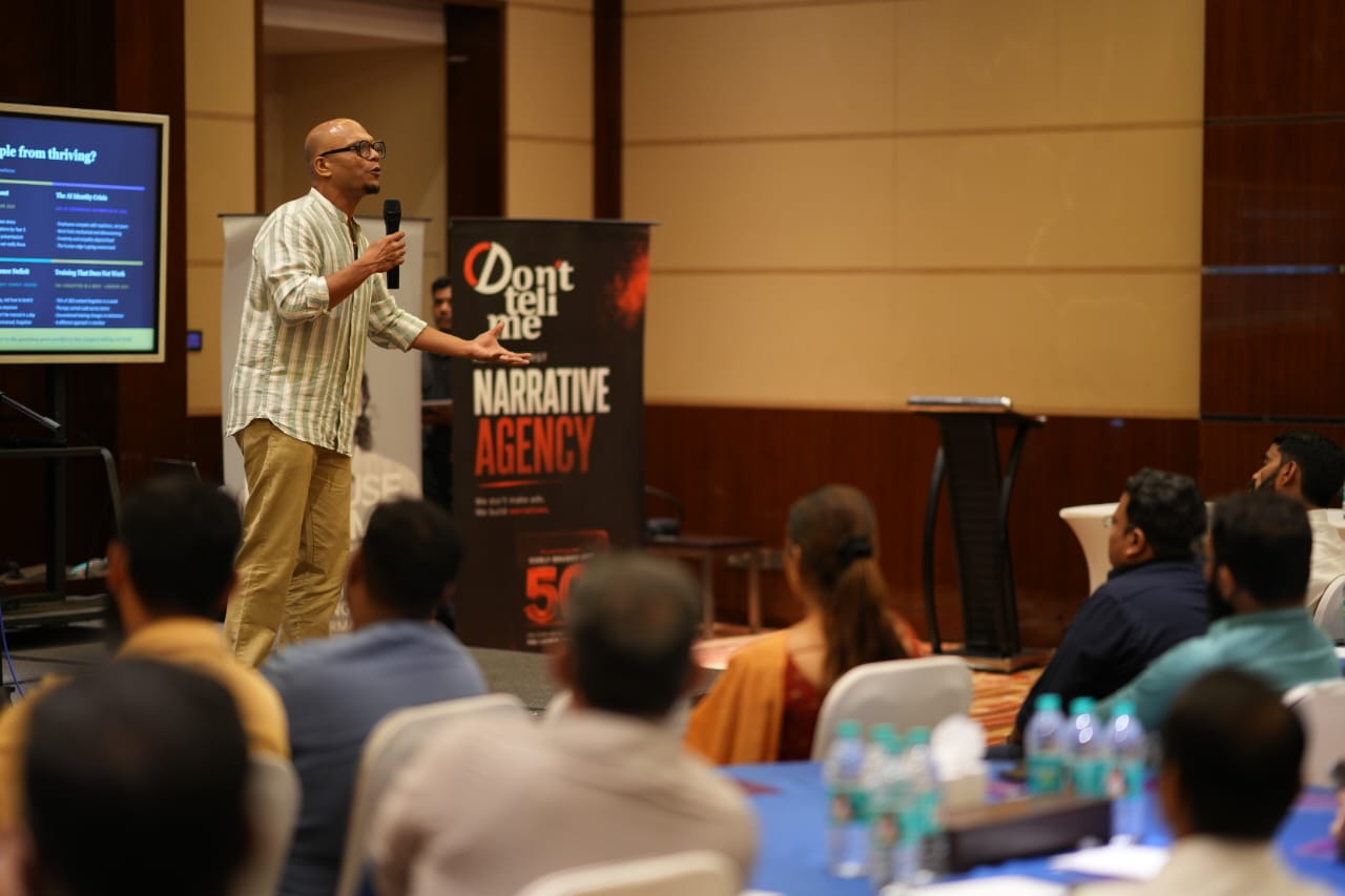 Saji Oorali addressing an audience seated at round tables in a conference room, a presentation slide behind him.