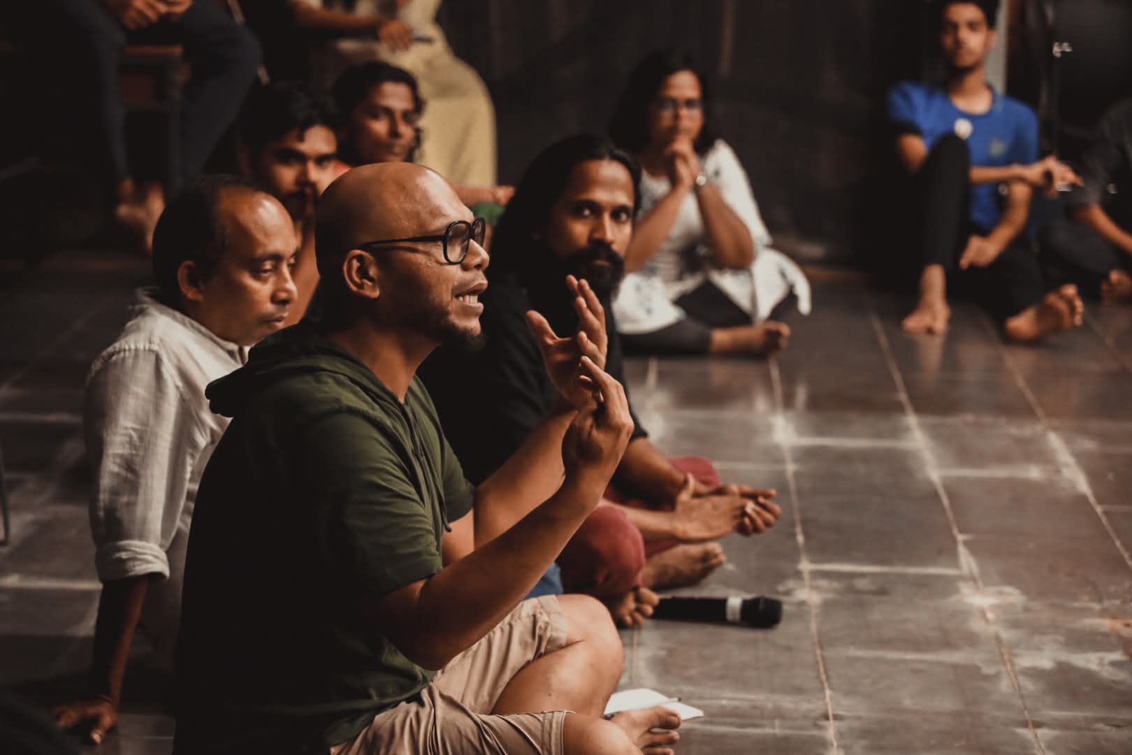 Saji Oorali sitting on the floor mid sentence, gesturing, with participants seated in a circle around him.