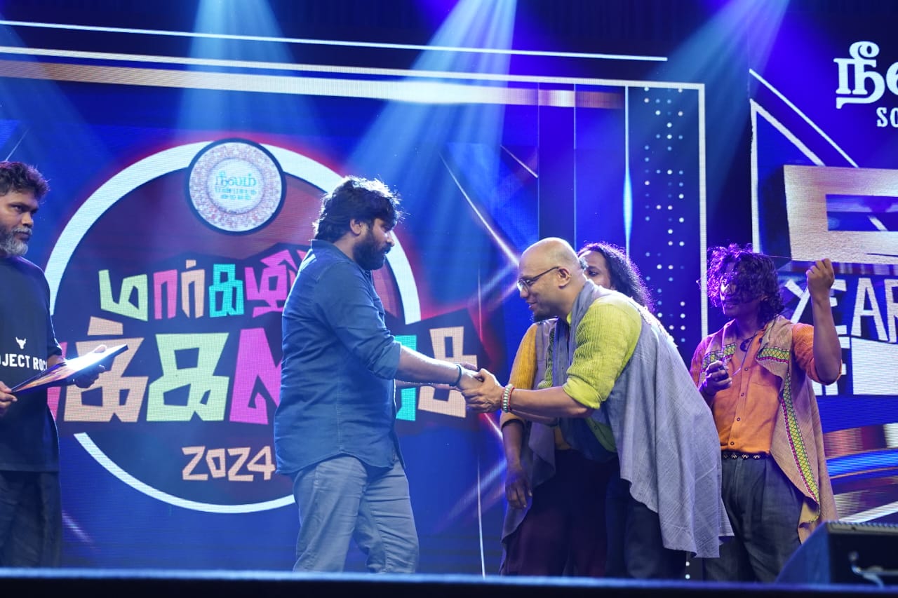Saji Oorali shaking hands on stage at a music awards event alongside other musicians.