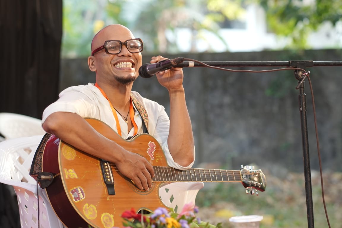Saji Oorali smiling broadly while singing into a microphone, playing a painted acoustic guitar outdoors.