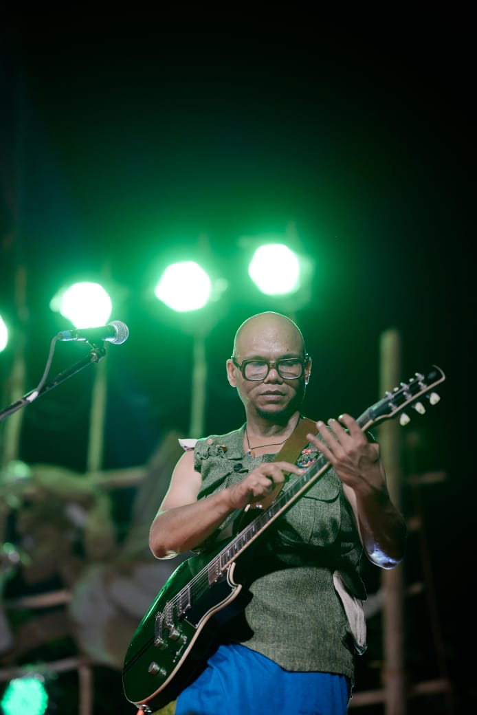 Saji Oorali playing electric guitar on a darkened stage under green stage lights.